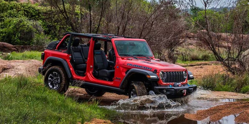 Red Jeep SUV driving through shallow water on an off-road trail surrounded by greenery