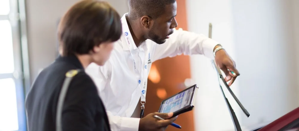A man in a white shirt with a name tag holds a tablet and points to the windshield wipers of a car.